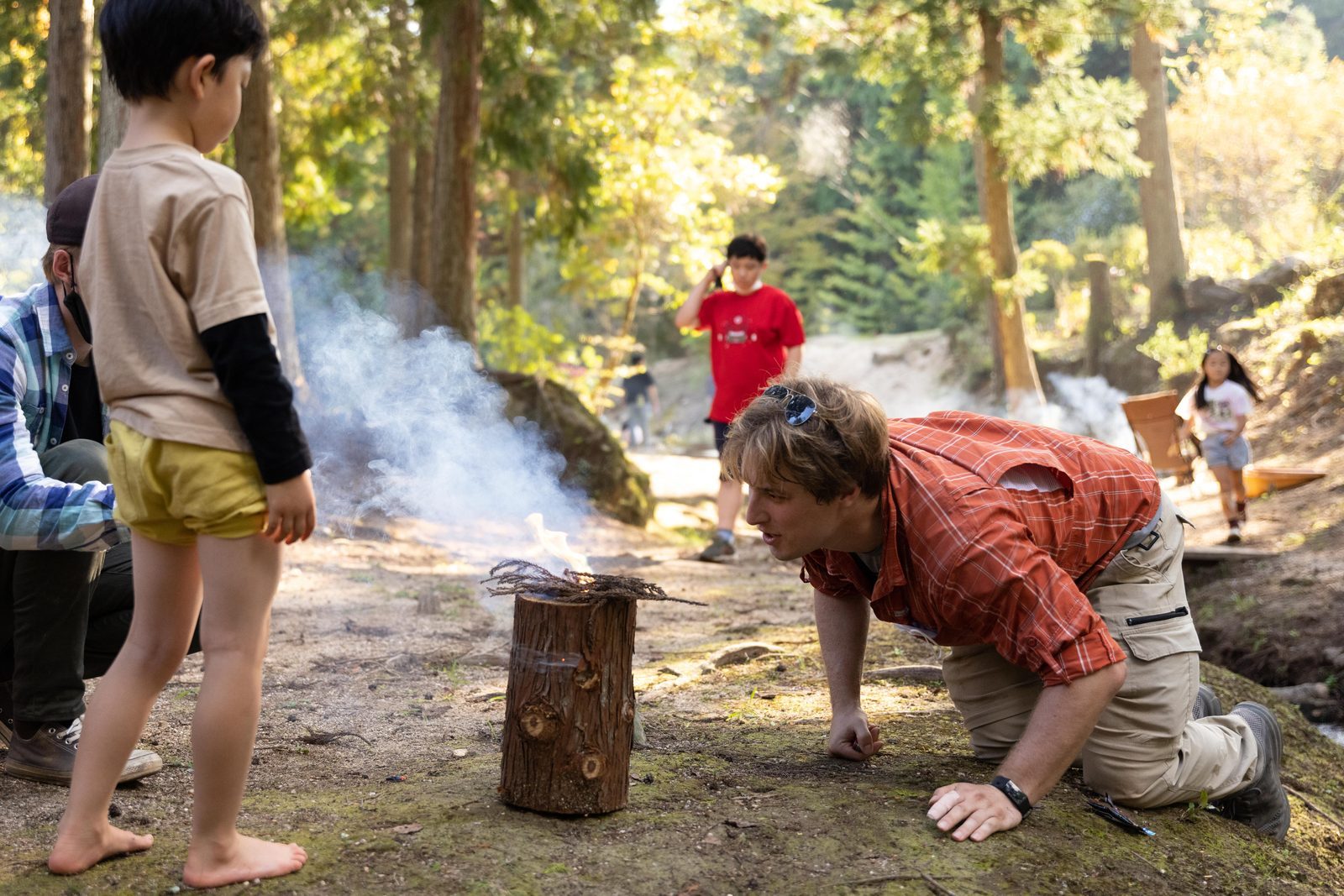 A guide kneeling to blow life into a fire while a child watches